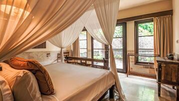 A bedroom with a four-poster bed, a wooden desk, and a view of the outdoors through large windows.