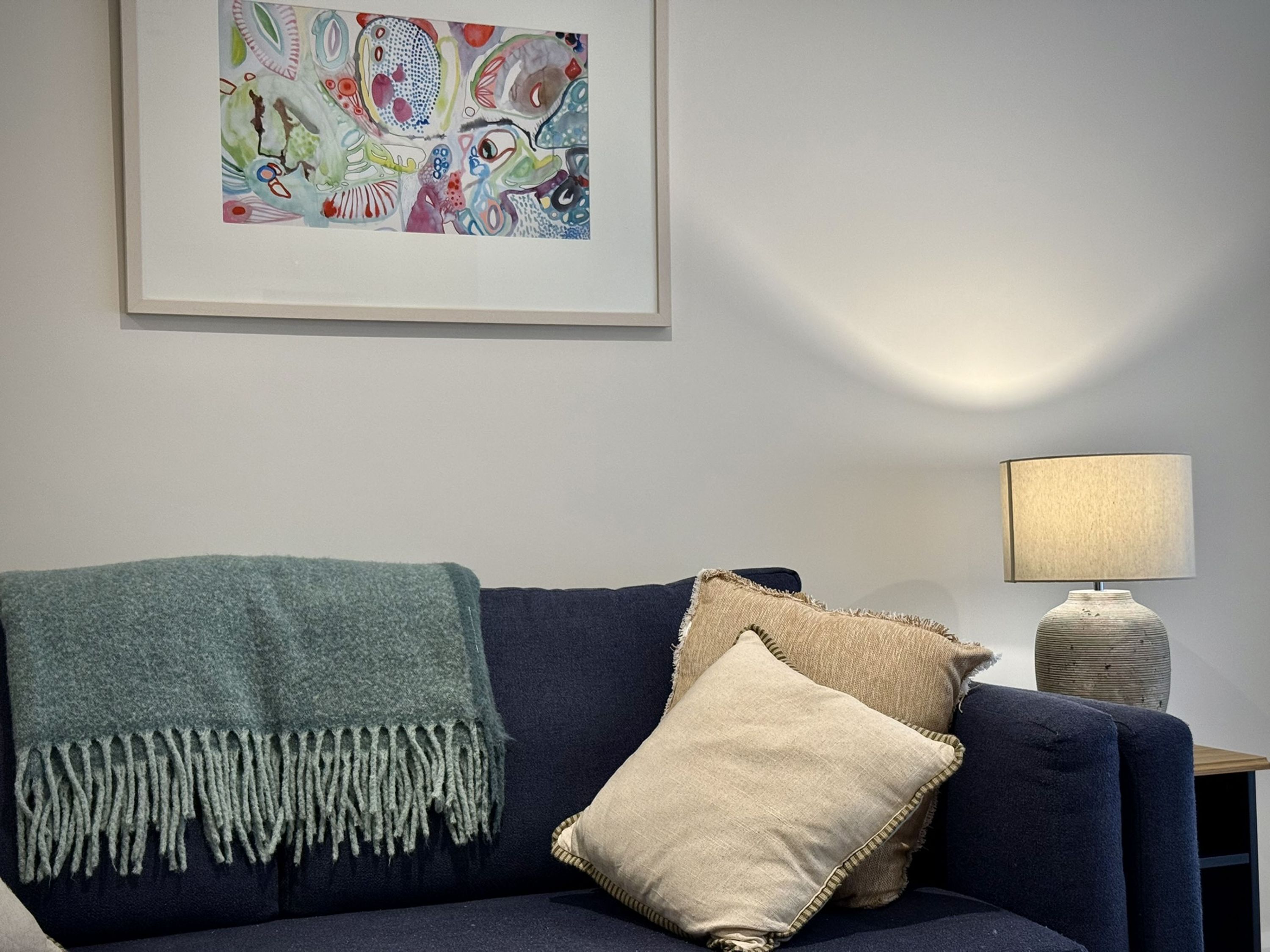 A dark blue sofa with a green throw blanket and two pillows, a lamp on a side table, and a colorful abstract painting on the wall.