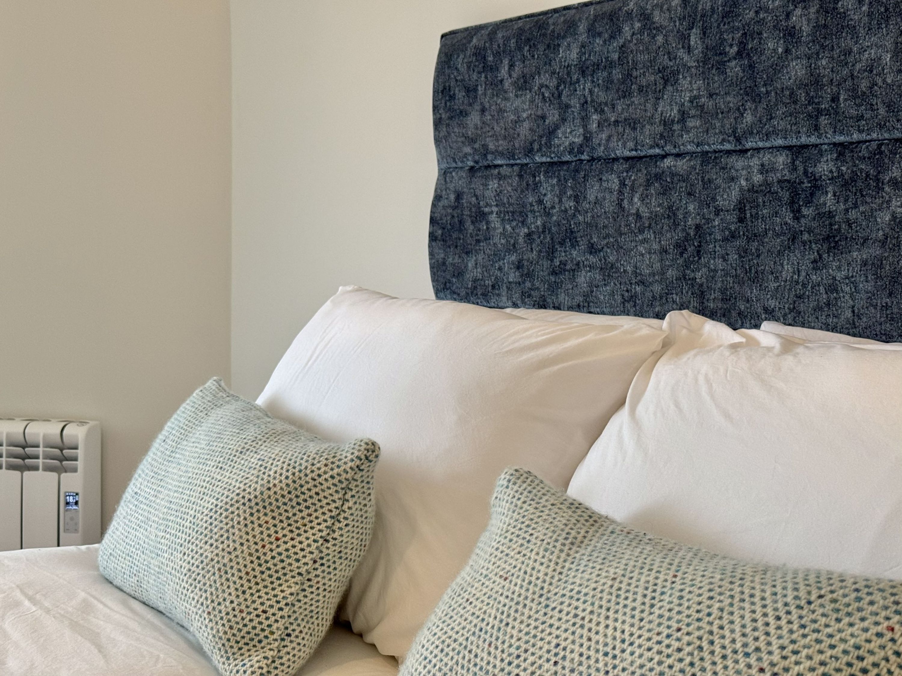 A bed with white bedding and two patterned pillows, a grey headboard, and a radiator in the background.