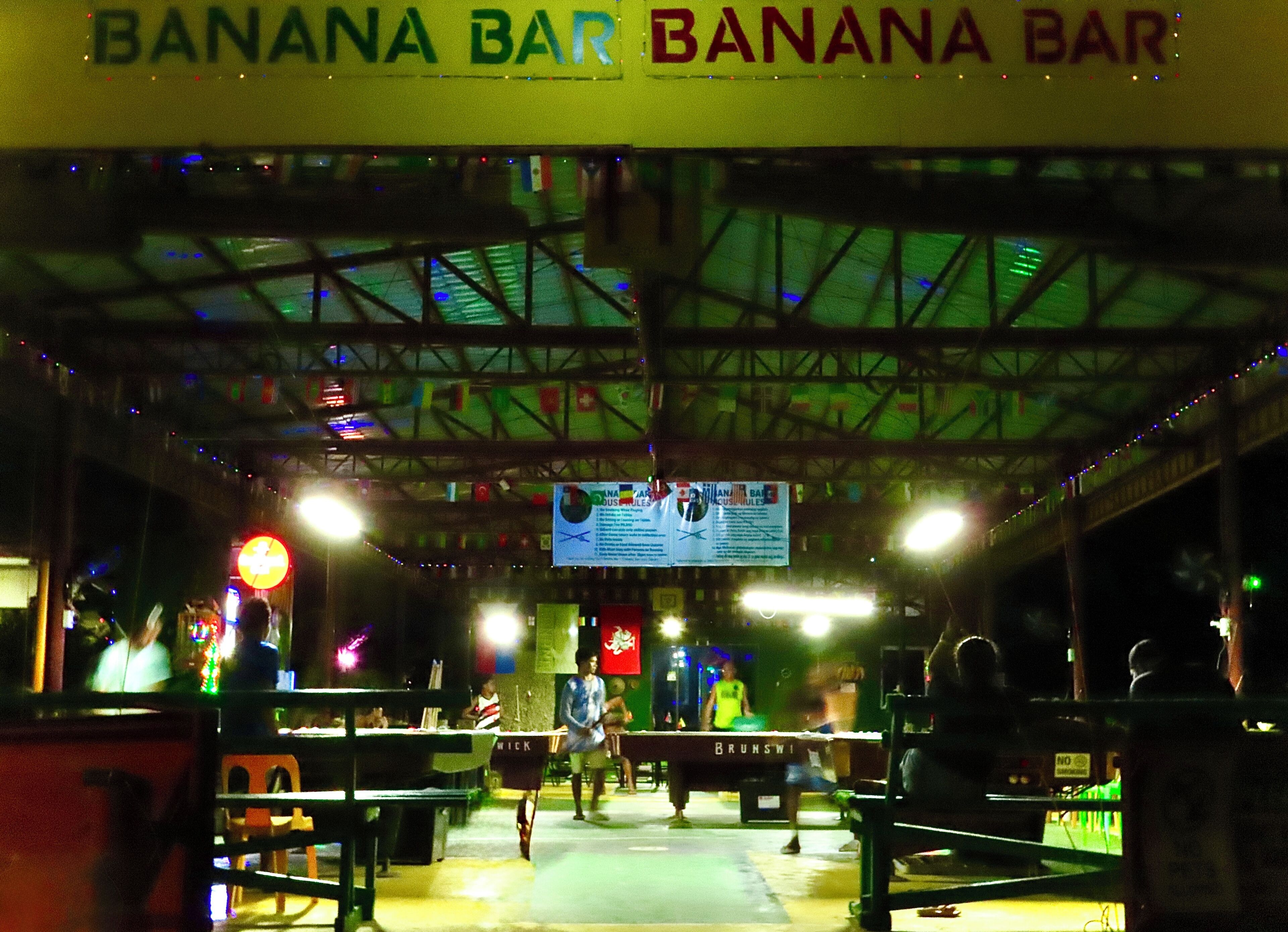 A Banana Bar with a high ceiling and metal beams, featuring tables and chairs for patrons.