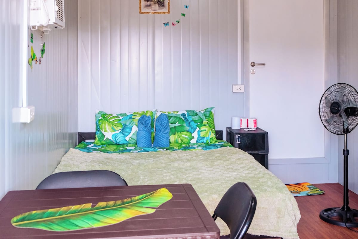 A room with a bed, a table with chairs, a standing fan, and a white door.