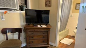 Une pièce avec un téléviseur posé sur un meuble en bois, une chaise et une salle de bain visible à travers une porte ouverte.