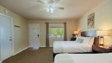 A hotel room with two beds, a ceiling fan, a window with blinds, a door, and a wall-mounted lamp.