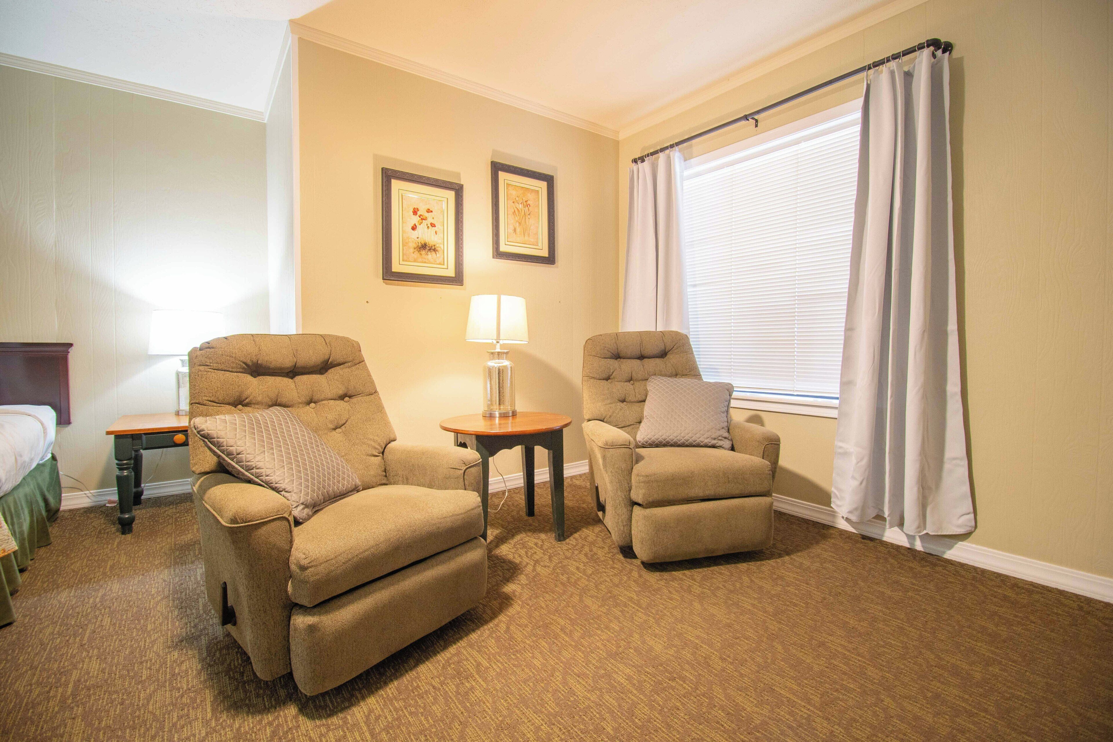 A hotel room with two armchairs, a small table with a lamp, a bed, and a window with curtains.