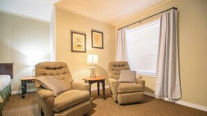 A hotel room with two armchairs, a small table with a lamp, a bed, and a window with curtains.
