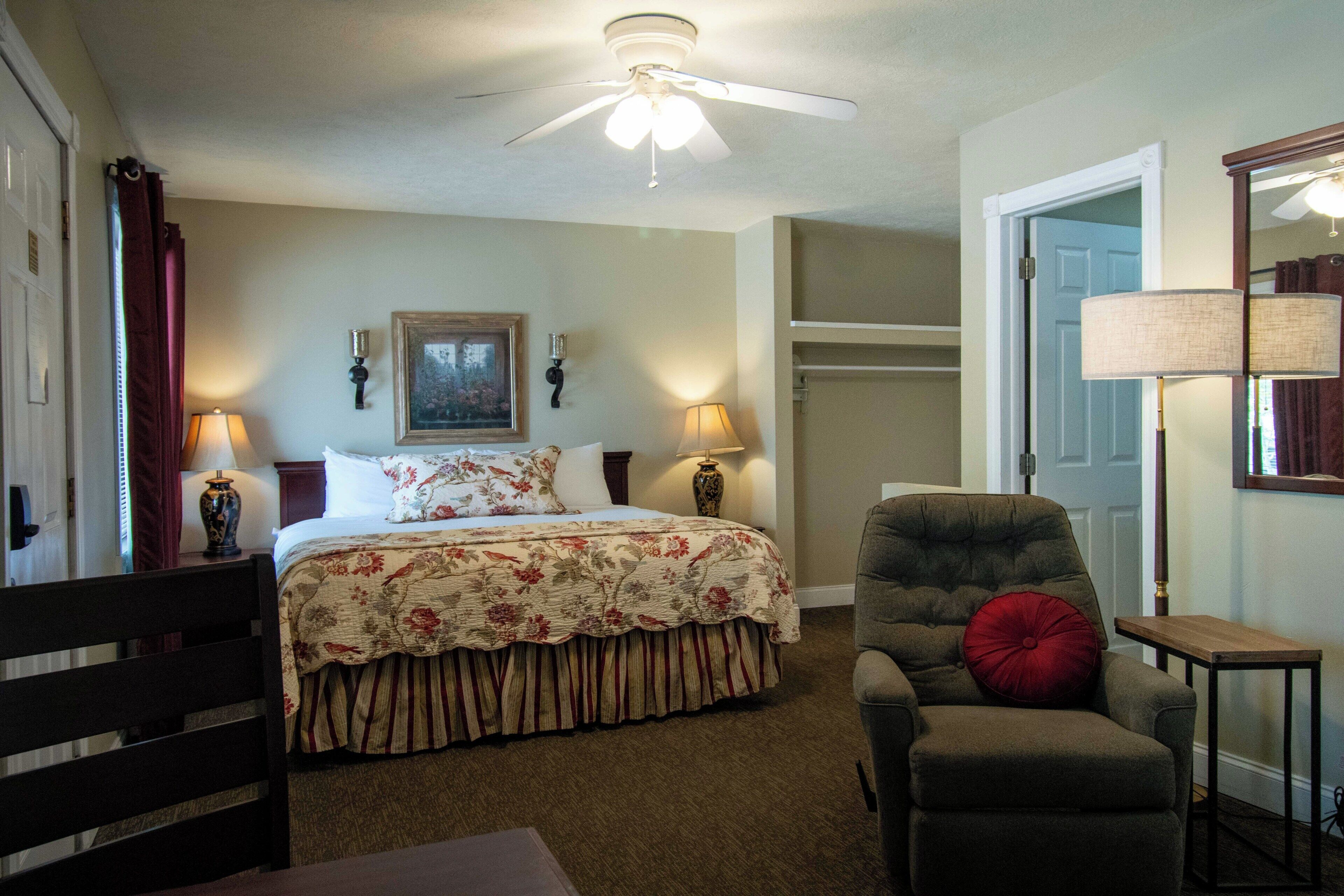 A bedroom with a bed, a chair, a ceiling fan, a lamp, and a door.