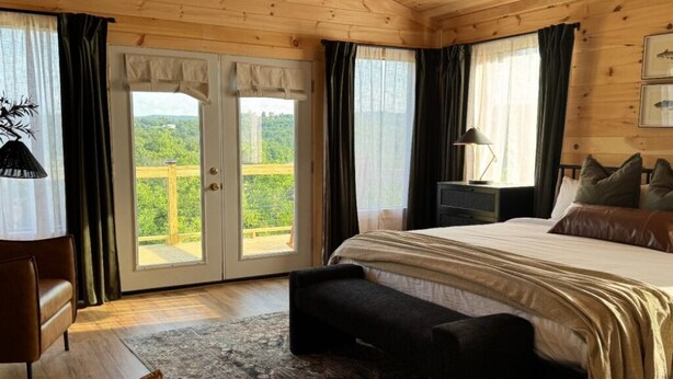 Un dormitorio con cama, banca, mesita de noche, lámpara y vista a follaje por una puerta.