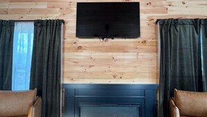 Una habitación con chimenea, sillones de cuero, televisor de pantalla plana montado en una pared de madera y cortinas.