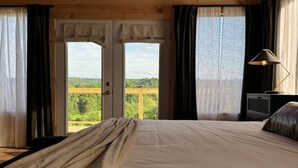 Un dormitorio con cama, cortinas, televisor y vista a la vegetación a través de una puerta.