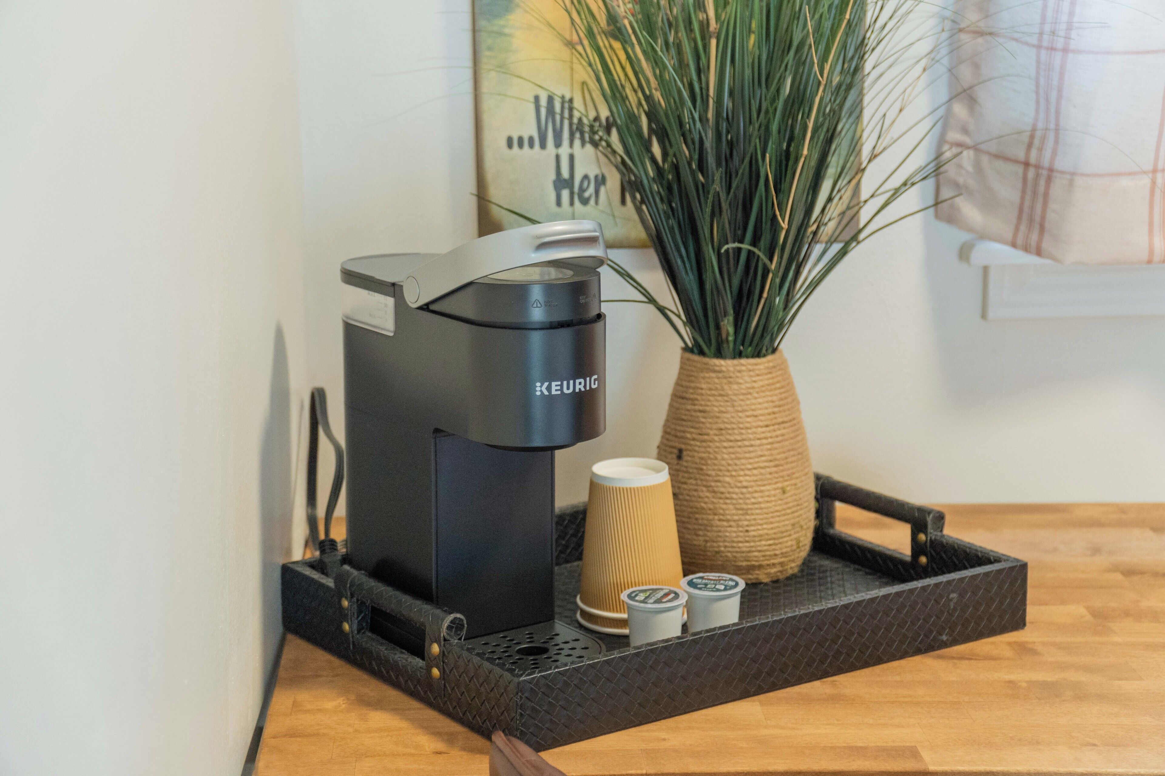 Una cafetera Keurig sobre una bandeja con un vaso de papel y dos cápsulas, una cesta trenzada con plantas verdes y una pared con un cartel.