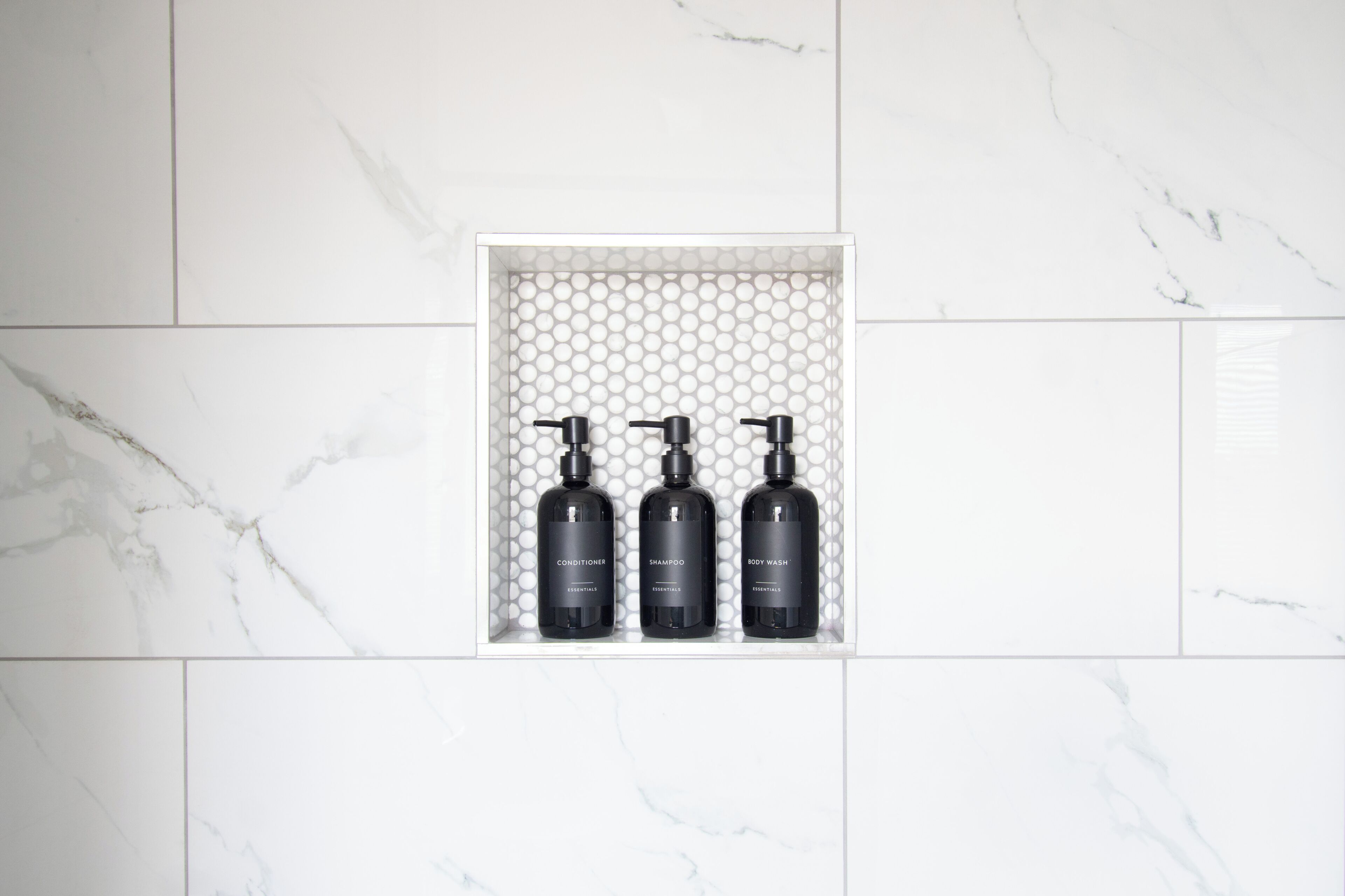 A bathroom with a marble wall and a shelf containing three black bottles labeled conditioner, shampoo, and body wash.