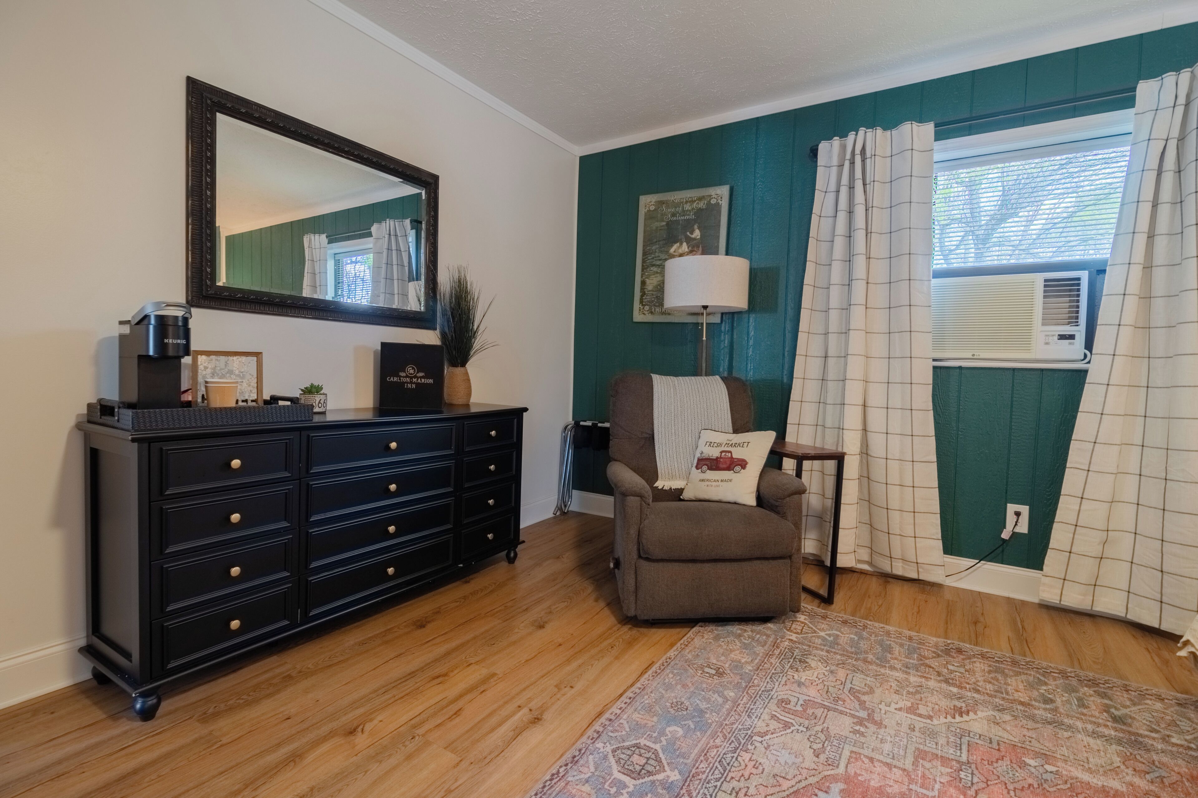 A room with a dark wooden dresser, a chair with a pillow, a rug, a mirror, and a window with curtains.