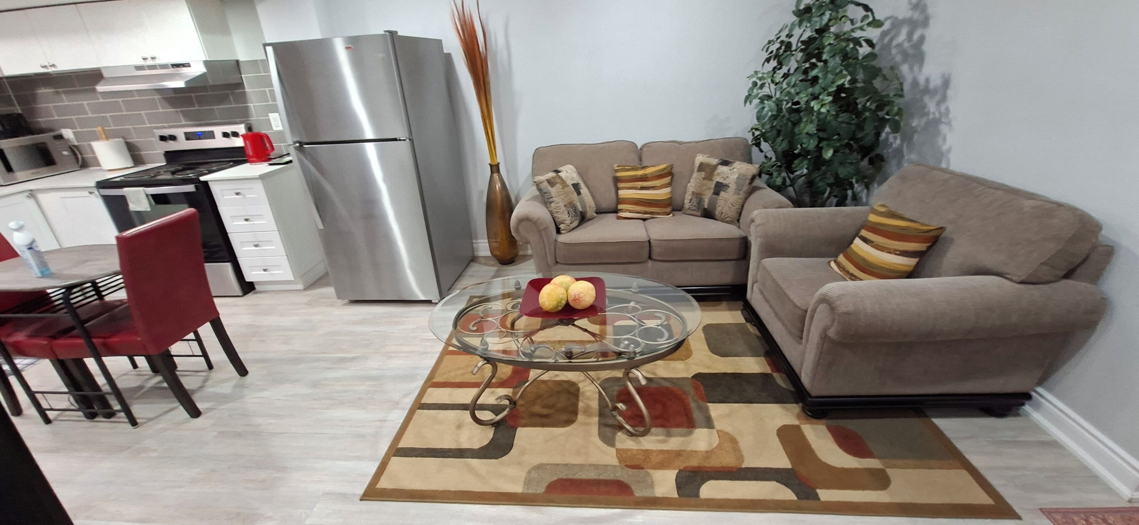 A modern kitchen with a stainless steel refrigerator, a dining area with a glass table and red chairs, and a beige sofa with patterned cushions.
