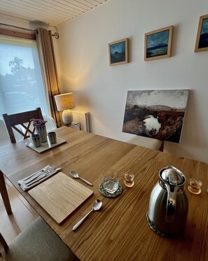 A dining table with a set of cutlery, a glass, a teapot, and a lamp. There are two framed pictures on the wall. - Two Miles From Town (Inverness)