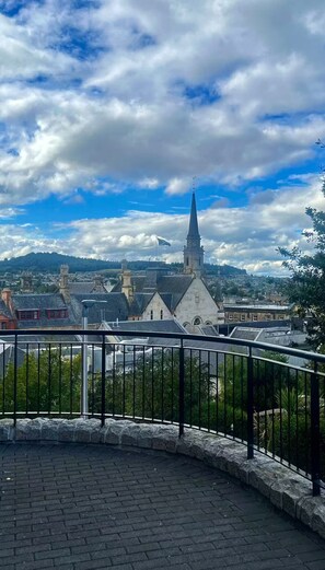 A view of a town with a prominent church tower, a paved area with a metal railing, and a distant hill. - Two Miles From Town (Inverness)