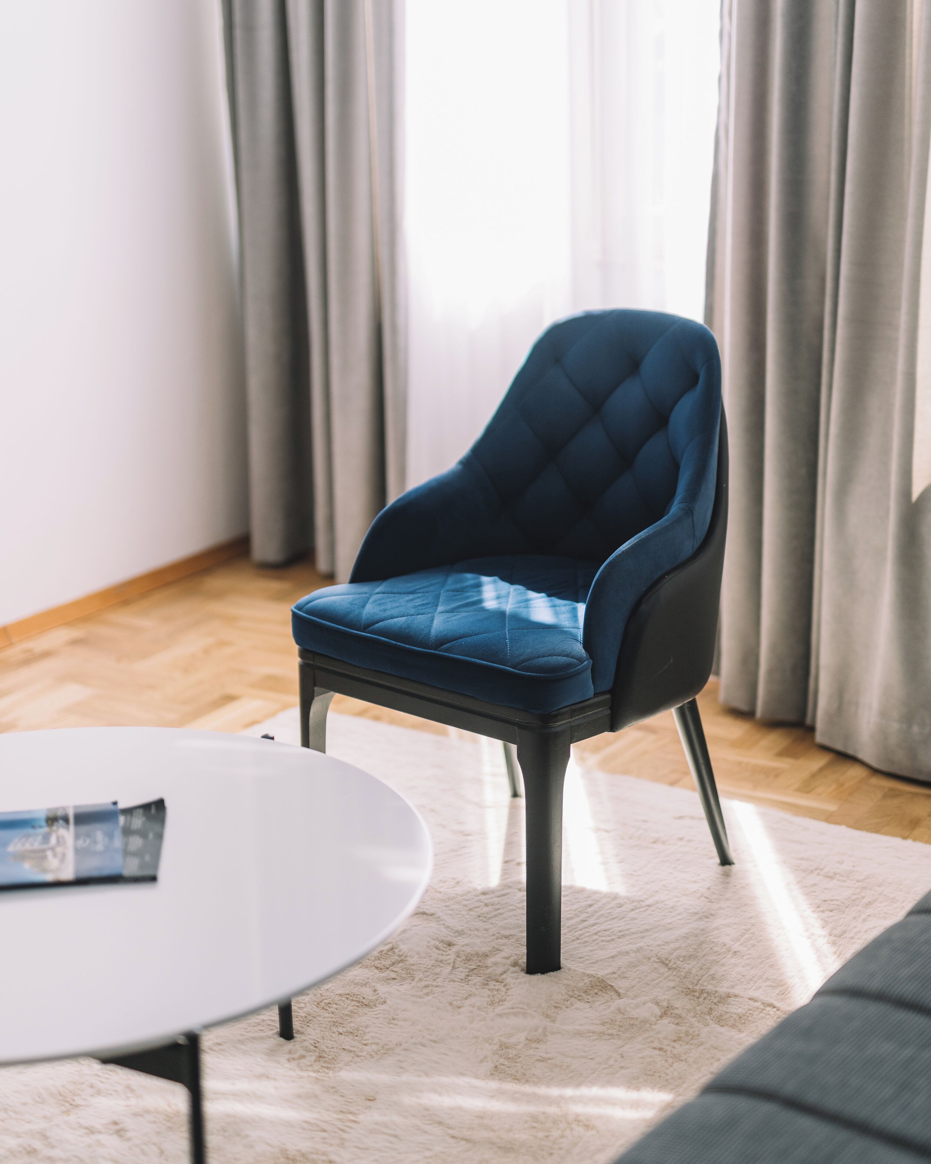 Ein modernes Zimmer mit einem dunkelblauen Polsterstuhl, einem weißen runden Tisch und einem hellen Teppich.