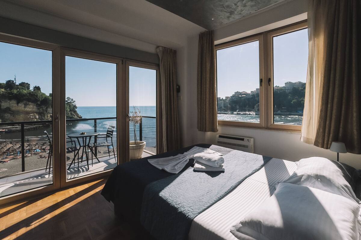 Habitación de hotel con cama, ventana con vista al mar y balcón con mobiliario exterior.