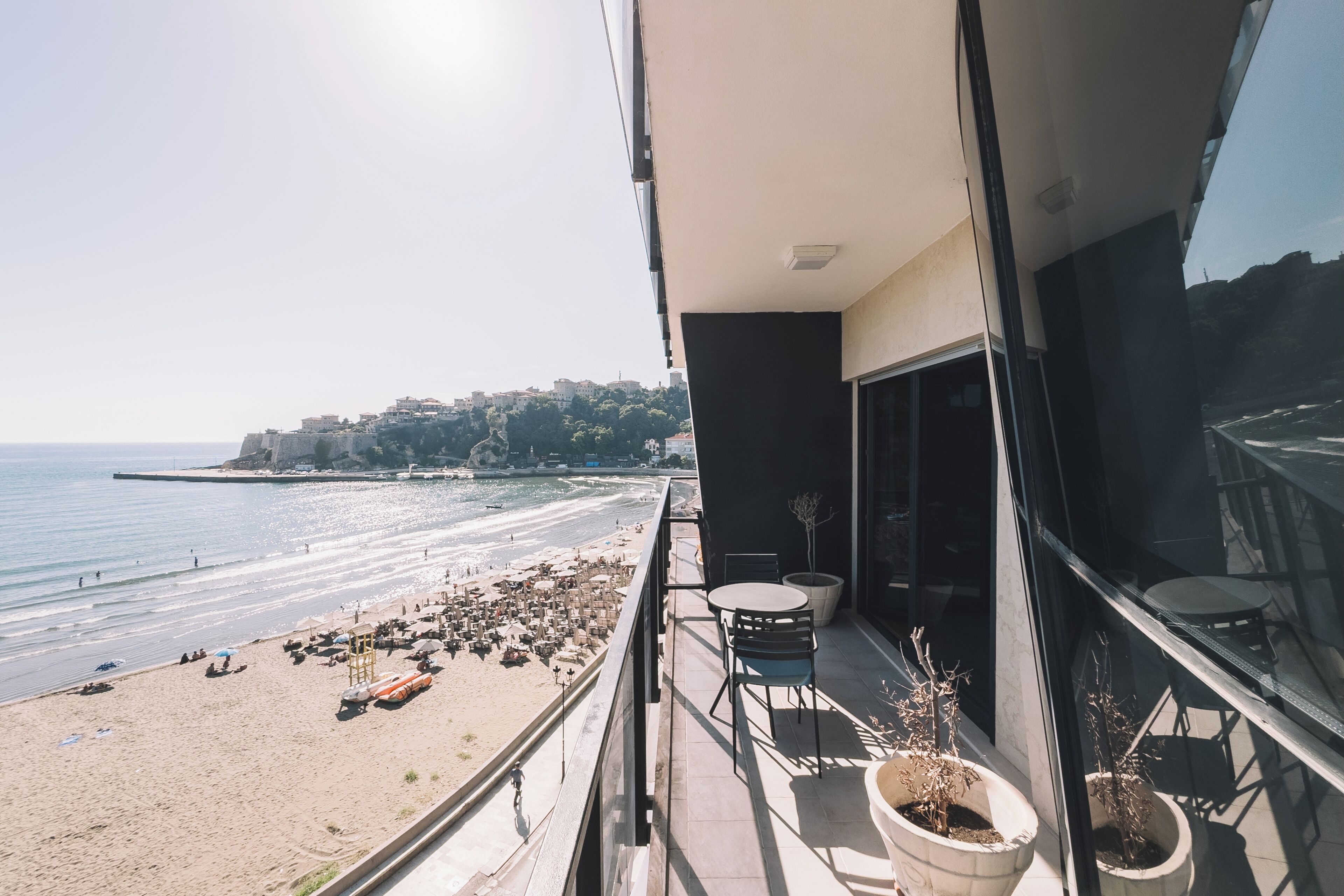 A balcony with a view of a beach, a dining area with chairs, and potted plants.