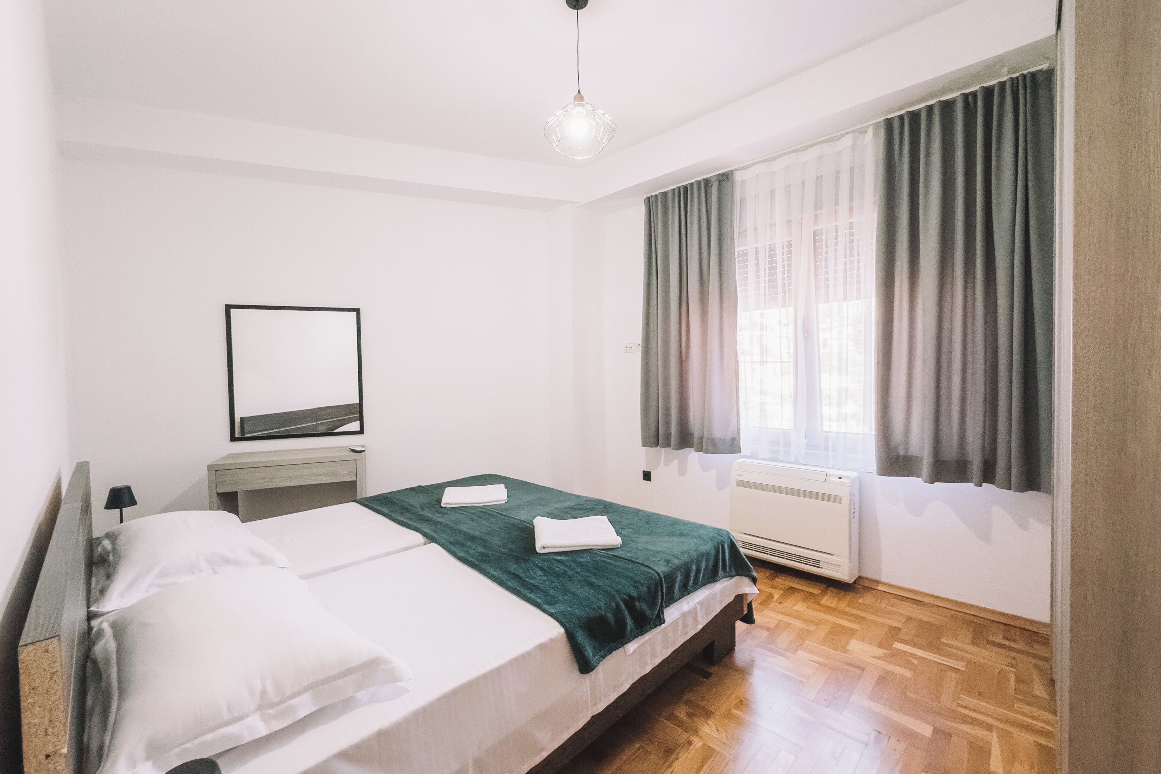 A bedroom with a bed, a bedside table, a mirror, a window with curtains, and a radiator.