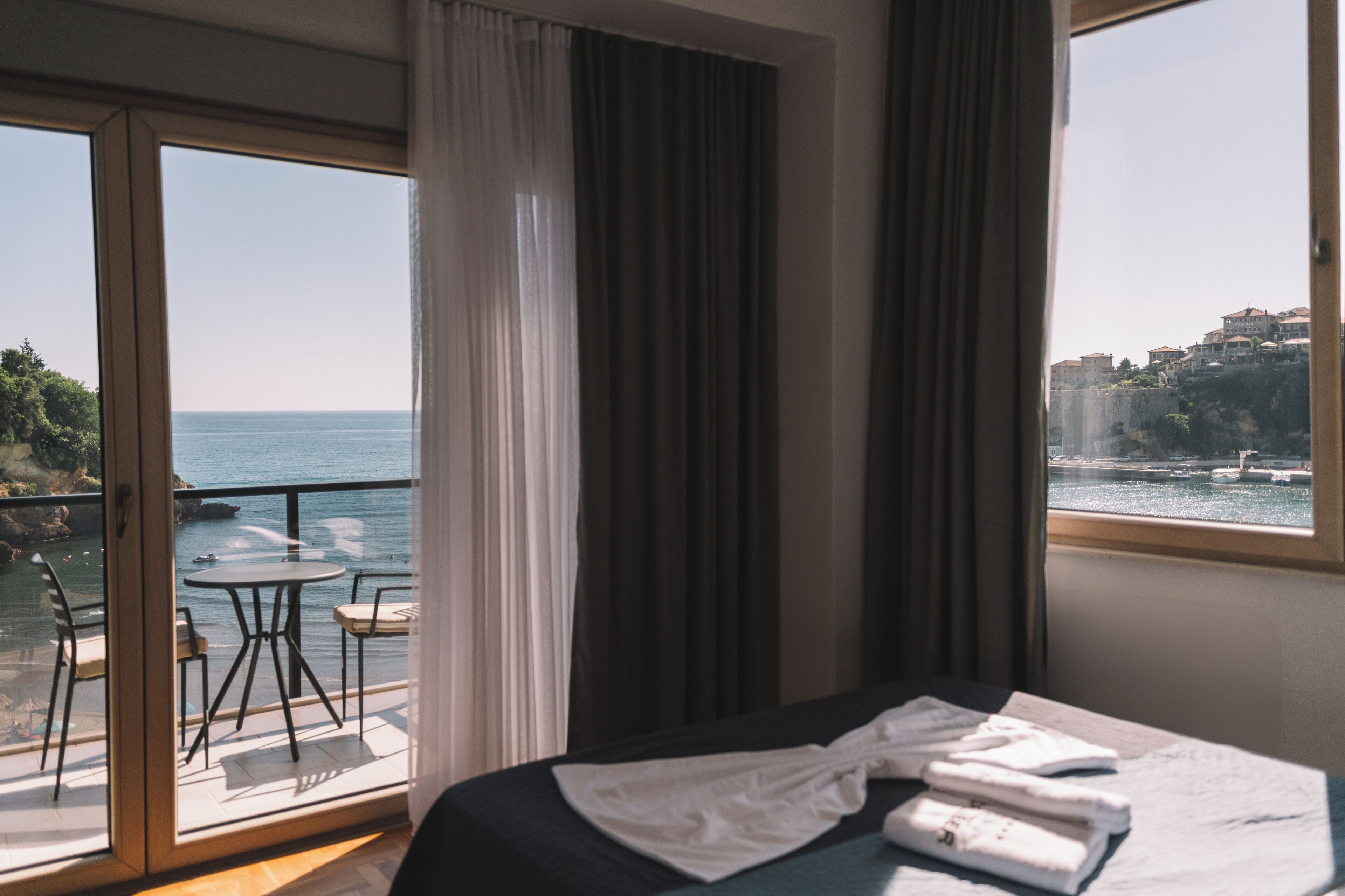 A hotel room with a bed, a table, and chairs on a balcony with a view of the sea and buildings.
