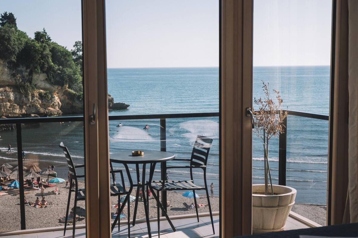 Una habitación con cama, una mesa y sillas, una planta en maceta y vista al mar a través de un ventanal.