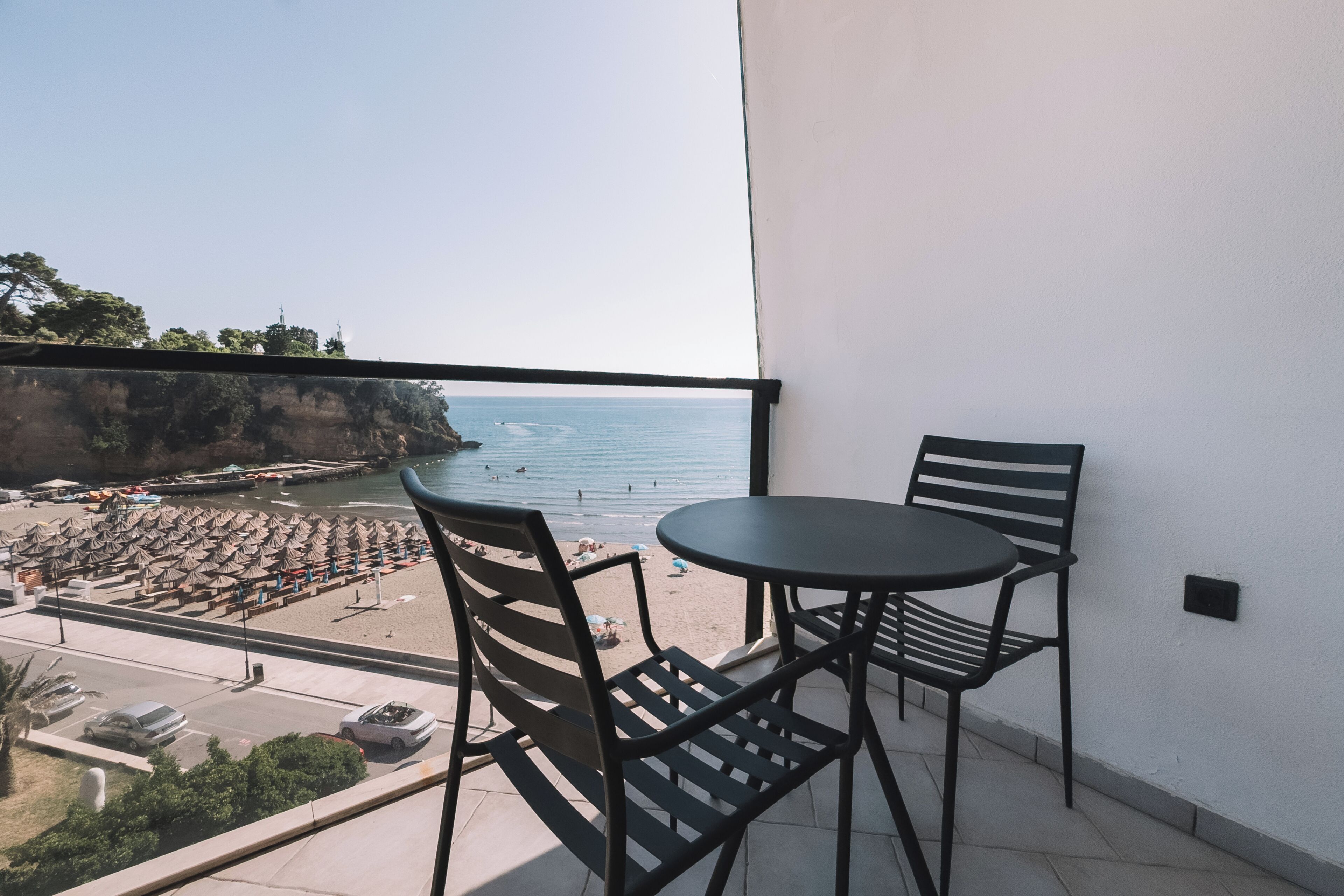 Un balcón con dos sillas y una mesa, con vista a una playa con sombrillas y una carretera.