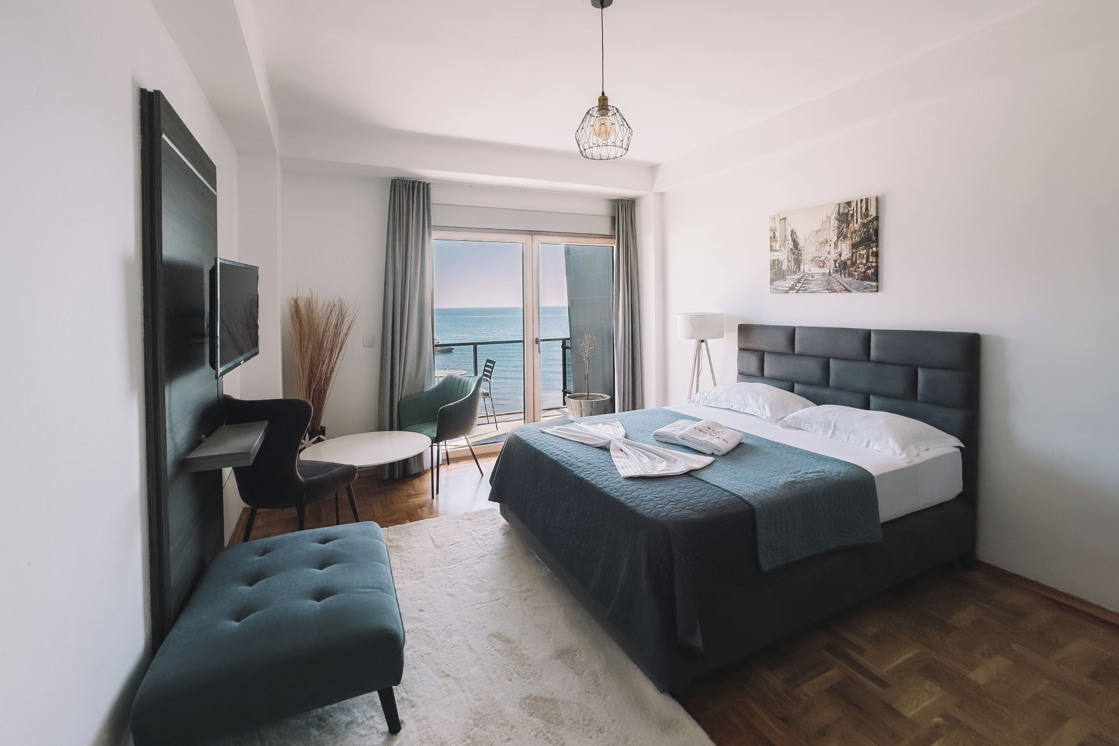 A modern hotel room with a bed, a desk, a chair, a bench, a TV, a lamp, and a view of the sea.