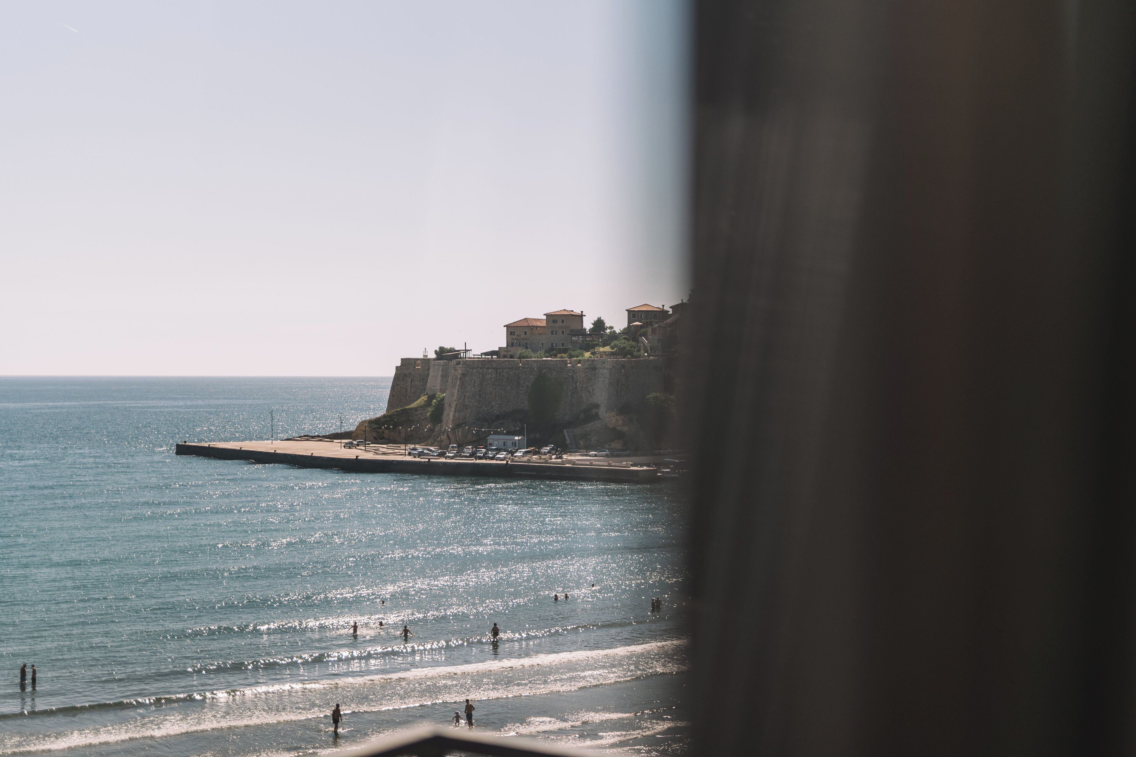 A coastal view with buildings on a cliff, a pier, and a beach with people.