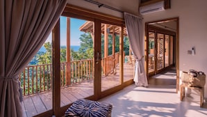 A room with a large sliding glass door leading to a balcony, wooden flooring, a zebra-patterned ottoman, and a wicker basket on a small table. - Organic Luxury Villa with Infinity Pool & Ocean Views (Karangasem)