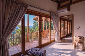 A room with a large sliding glass door leading to a balcony, wooden flooring, a zebra-patterned ottoman, and a wicker basket on a small table. - Organic Luxury Villa with Infinity Pool & Ocean Views (Karangasem)