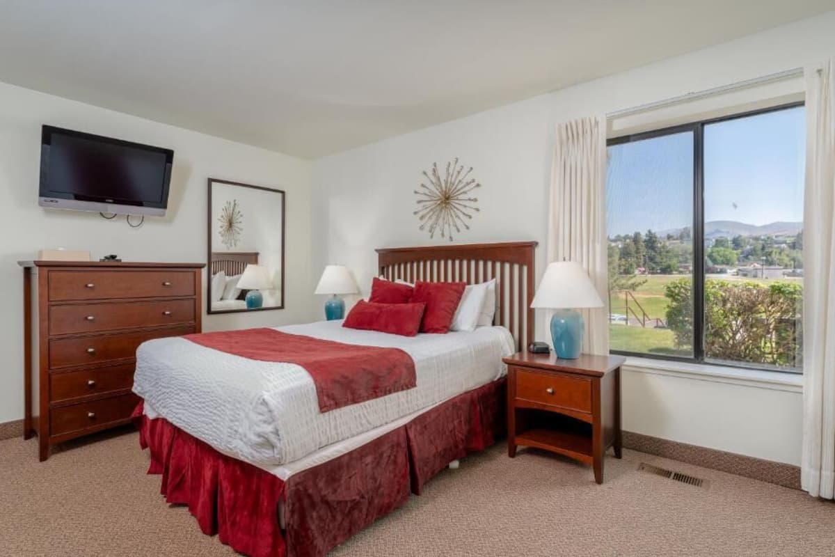 Habitación de hotel con cama, televisor, cómoda, mesita de noche y ventana con vista.