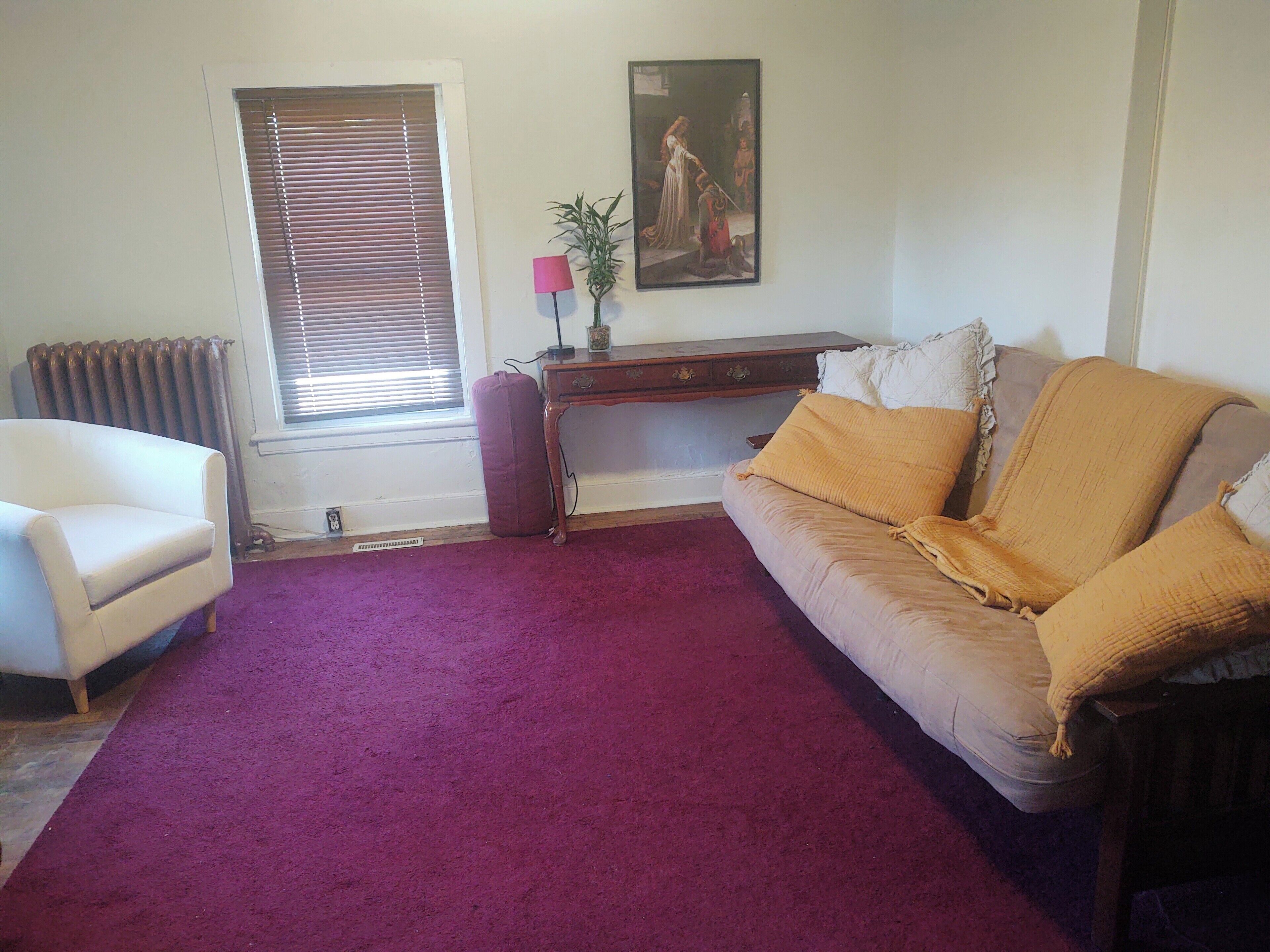 A living room with a sofa, a chair, a radiator, a window with blinds, a desk with a lamp, and a painting on the wall.