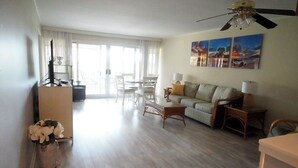 Un salon spacieux comprenant un canapé, une table basse et un meuble pour la télévision. On y trouve également un ventilateur de plafond, un coin repas avec des chaises et une grande fenêtre avec des rideaux.