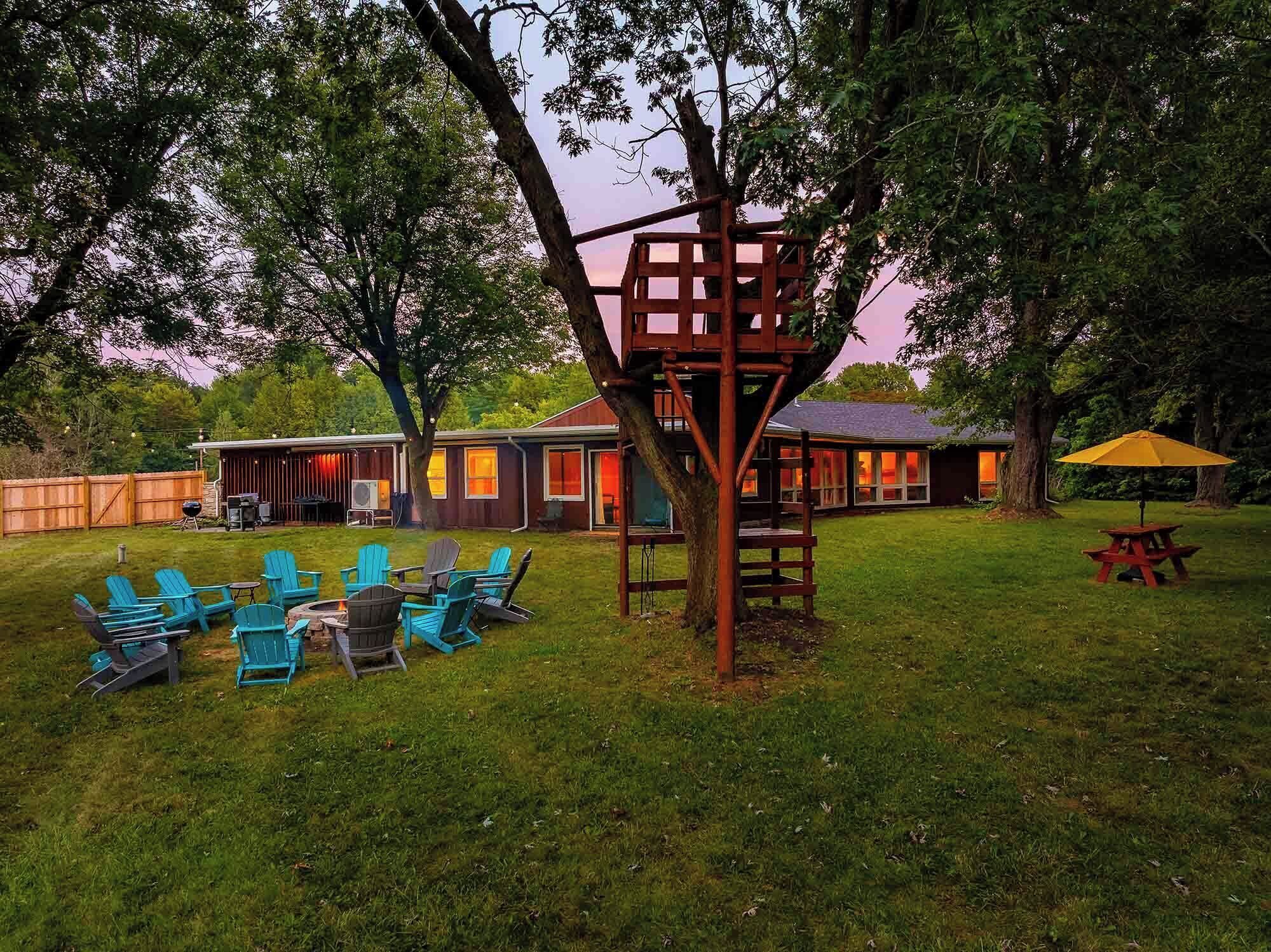 A backyard with a fire pit, outdoor seating, and a treehouse structure.