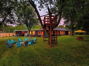 A backyard with a fire pit, outdoor seating, and a treehouse structure.