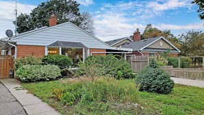Uma casa residencial com telhado inclinado, chaminé de tijolos e um jardim bem cuidado com diversas plantas e cerca de madeira.