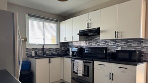 A modern kitchen with white cabinets, a black countertop, a stainless steel oven, a microwave, and a window with blinds.