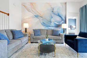 Ein Wohnzimmer mit grauem Sofa, einem blauen Sessel, einem Glastisch und einem großen abstrakten Gemälde an der Wand.