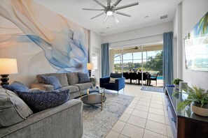 Ein modernes Wohnzimmer mit Sofa, Sessel und Couchtisch, einem Deckenventilator und einem großen Fenster mit Blick ins Freie.