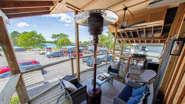 Overdekt terras met rieten meubilair, een centrale verwarmingsunit en uitzicht op een parkeerplaats en een meer.