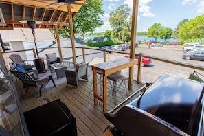 A covered deck with outdoor furniture, a grill, and a view of a parking lot and lake.