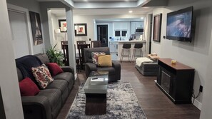 Un salon avec un canapé, des fauteuils, une table basse et un téléviseur. On aperçoit une cuisine au fond.