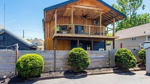 Een houten huis met twee verdiepingen, een balkon, een plafondventilator en een omheinde tuin met drie struiken.