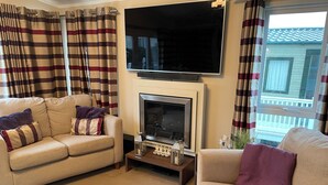 A living room with a sofa, a fireplace, a flat-screen TV, and a window with curtains. - Iris Lodge, 3 bedroom, dogs stay free at Rockley Park, Poole Harbour, Dorset (Poole)