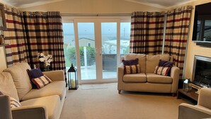 A living room with two beige sofas, a coffee table, a fireplace, and a view through a glass door. - Iris Lodge, 3 bedroom, dogs stay free at Rockley Park, Poole Harbour, Dorset (Poole)