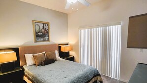 Een slaapkamer met een bed, twee nachtkastjes met lampen, een plafondventilator, een raam met jaloezieën en een aan de muur bevestigde televisie.