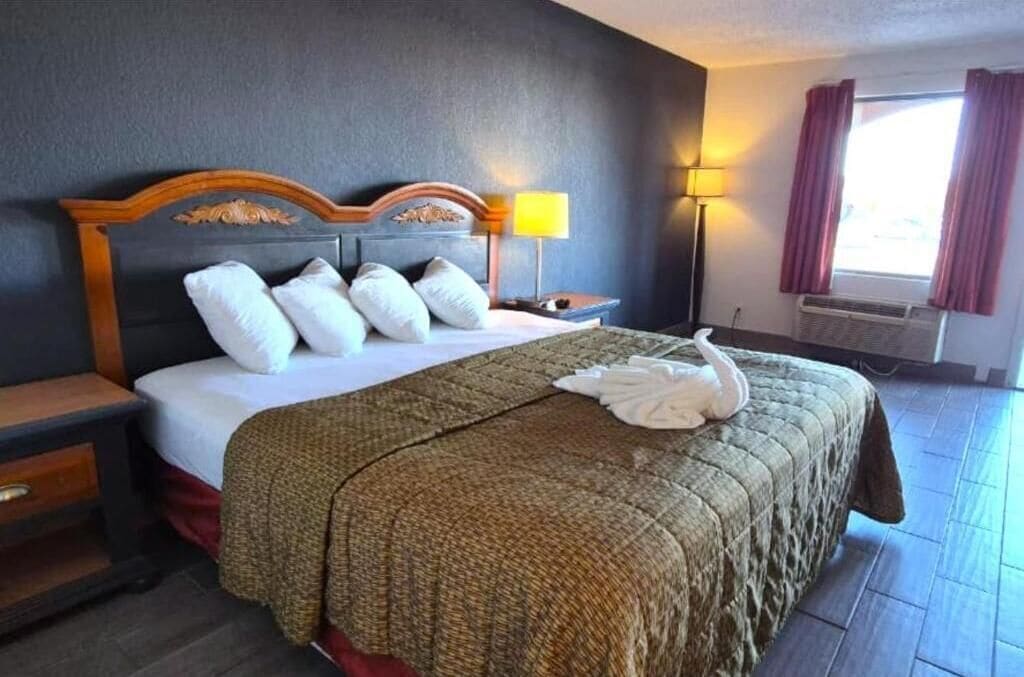 A hotel room with a bed, a nightstand, a lamp, a window with curtains, and a wall-mounted air conditioner.