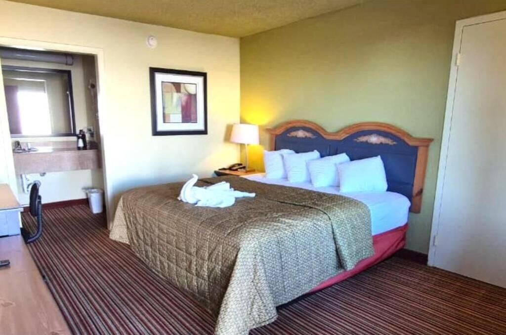 Habitación de hotel con cama, mesita de noche con lámpara, un cuadro enmarcado en la pared y un baño visible a través de una puerta abierta.