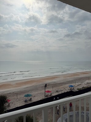 Blick auf einen Strand von einem Balkon mit Geländer, feiner Sand und einige Sonnenschirme.