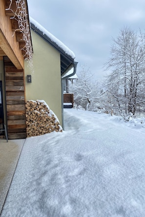Ein modernes Haus mit Schnee bedeckter Fassade, einer Holztreppe, einem Holzstapel und einem verschneiten Baum im Hintergrund.